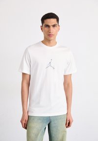 Valge puuvillane t-särk, millel on keskel hall Jordan logo, lühikesed varrukad ja klassikaline ümar kaelus, koos helesiniste denim teksadega.