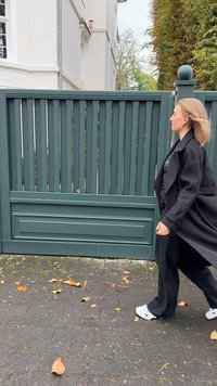 Fekete hosszú kabát, fekete ing és bővített nadrág. Világosszürke sportcipő. Zöld, függőleges, lamellás kerítés és lehullott őszi levelek a háttérben.