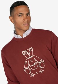 Bordeauxroter Sweatshirt mit Rundhalsausschnitt, versehen mit einem weißen gestickten Teddy-Bären-Design und dem Text "Harper & Neuer" darunter.