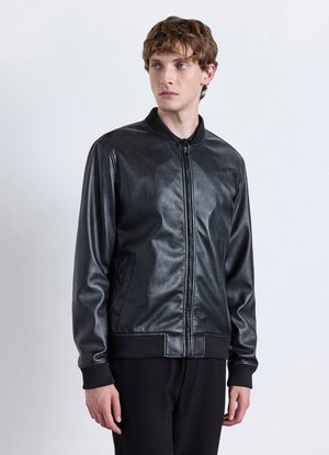 Junger Mann, der eine schwarze Lederjacke mit Reißverschluss und schwarze Hose trägt, steht und schaut leicht nach rechts vor einem schlichten Hintergrund.