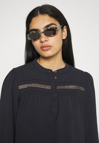 Blusa preta de manga longa com decote redondo, fecho de botão e detalhes em renda na zona do peito. Modelo a usar óculos de sol cinzentos com padrão.