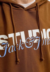 Hnědá mikina s kapucí, na které je velký bílý a světle modrý text s nápisem "STUDIO Jack & Jones." Text je tučný a obsahuje dekorativní prvky.