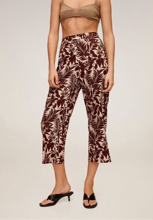 Vrouw die cropped bruine en beige bloemenpatroon broek, een beige crop top en zwarte sandaal met hakken draagt, staand tegen een effen achtergrond.