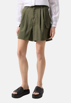 Kvinde iført olivengrønne plisserede shorts med sidelommer, hvid bluse med udskårne ærmer og sorte platform-slide sandaler.