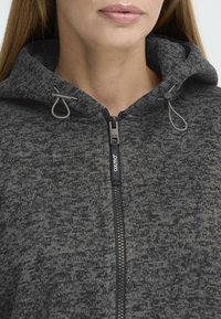 Donkergrijze zip-up hoodie van textuurstof, met verstelbare trekkoorden bij de capuchon en een prominent logo op de ritssluiting.