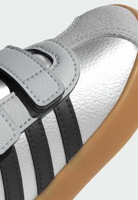 Strieborná detská topánka s čiernymi pruhmi, dvoma šedými suchými zipsami a béžovou gumovou podrážkou, zobrazená v detailnom zábere.