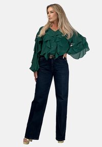 Zielona, falbaniasta bluzka z długimi rękawami, w połączeniu z ciemnoniebieskimi dżinsami o wysokim stanie. Posiada złotą klamrę paska i jasne buty na platformie.