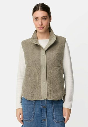 Kobieta ubrana w beżową kamizelkę polarową zapinaną na guziki, na białej koszuli z długim rękawem i niebieską spódnicę dżinsową, stojąca na jednolitym tle.