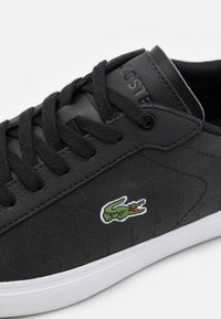 Baskets noires avec un dessus en cuir texturé, arborant un logo brodé vert et blanc, des lacets noirs et une semelle en caoutchouc blanche.