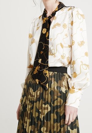 Mujer vestida con una blusa blanca con patrones de cadenas doradas, una bufanda negra con motivos dorados y una falda plisada de camuflaje.