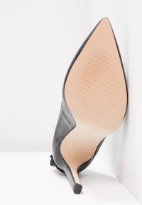 Chaussure à talon haut en cuir noir avec un bout pointu, un talon fin et une texture lisse. La semelle présente un motif rainuré pour une meilleure adhérence.