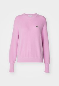 Pull rose en maille avec un col rond, des poignets et un ourlet côtelés. Présente un petit logo Lacoste vert sur le côté gauche de la poitrine.
