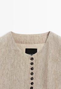 Beige linnen overhemd met ronde halslijn en een verticale rij van zeven zwarte knopen middenvoor.