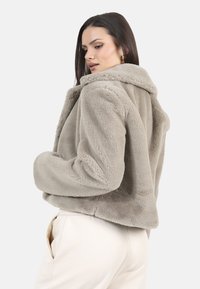 Kurtka z futra syntetycznego w jasnym taupe z kołnierzem, krótki fason i teksturowane wykończenie. W połączeniu z jasnymi, luźnymi spodniami.