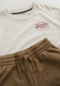 T-shirt vintage SuperDry écru à col rond avec logo rouge, associé à un pantalon marron à taille élastique avec cordon.