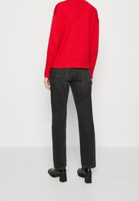 Personne debout de dos, portant un pull rouge, un jean noir à jambe droite et des bottines noires à talons sur un fond uni.