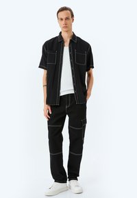 Camisa negra de manga corta con costuras blancas y dos bolsillos en el pecho, combinada con pantalones negros con detalles de parches y un bolsillo lateral. Zapatillas blancas.