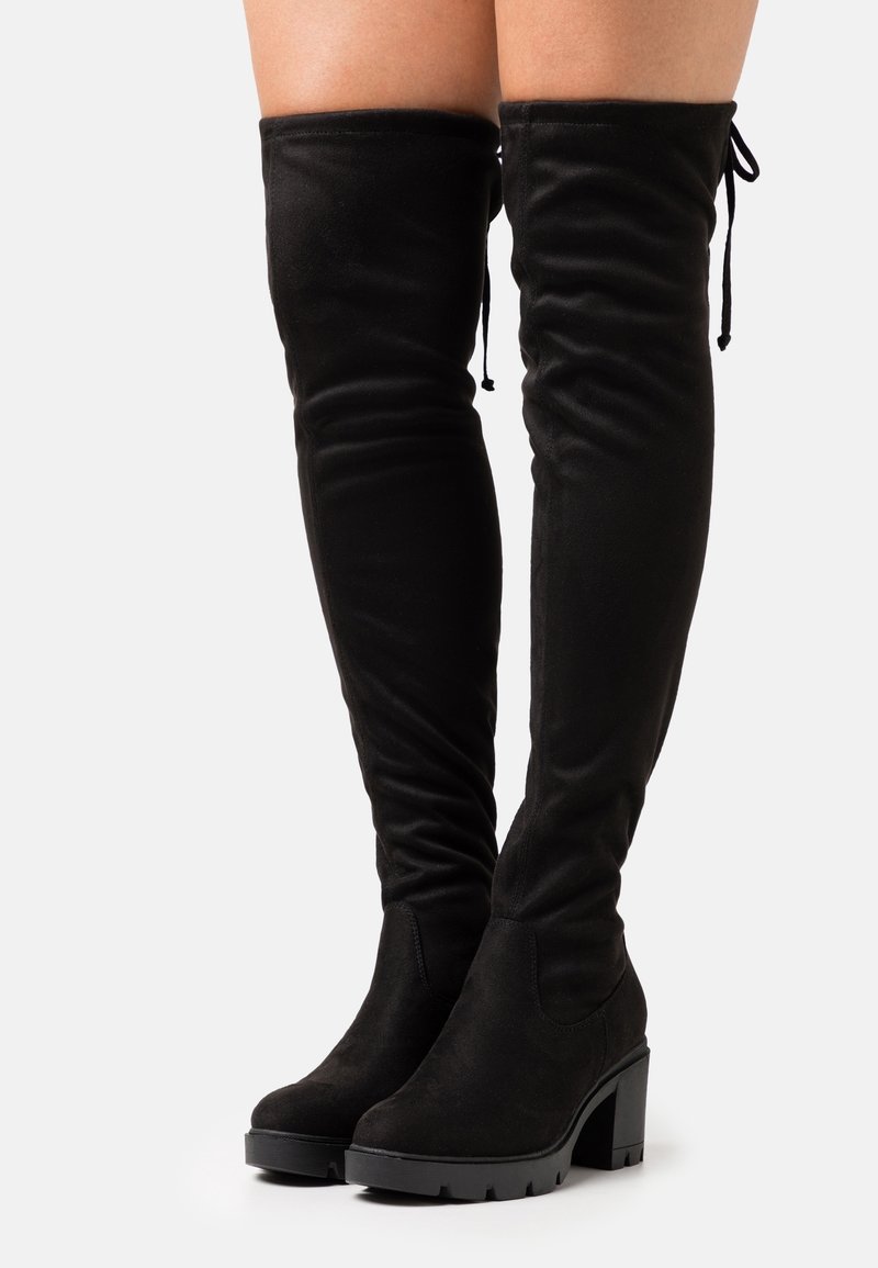 Bottes noires au-dessus du genou avec talons épais et semelles à crampons portées sur jambes nues, avec fermeture à nouer en haut à l'arrière.
