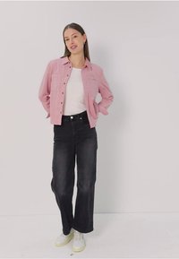 Rosa Button-Down-Hemd über einem weißen T-Shirt, kombiniert mit hoch taillierten schwarzen Jeans und weißen Sneakers mit gelben Akzenten. Heller Hintergrund.