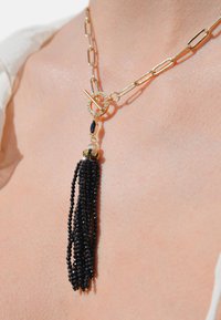 Collier en chaîne dorée avec un fermoir rond orné de pierres transparentes, un accent en perle noire et un long gland en perles noires.