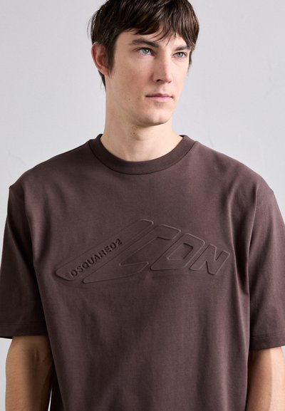 Camiseta de algodón marrón con cuello redondo, que presenta un diseño de logo en relieve en la parte frontal. Corte sencillo y relajado con mangas cortas.