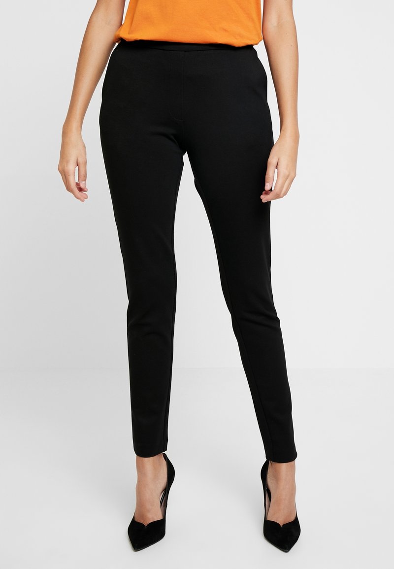 Femme portant un pantalon noir slim et des chaussures à talons noirs, associée à un haut orange, debout devant un fond clair uni.