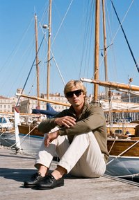 Homme portant des lunettes de soleil, une veste verte et un pantalon blanc, assis en tailleur sur un quai avec des voiliers et des bâtiments en arrière-plan.