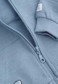 Jasnoniebieska kurtka zapinana na zamek z teksturowanej tkaniny i ściągaczami na rękawach. Posiada gładki zamek błyskawiczny oraz małą metkę na rękawie.
