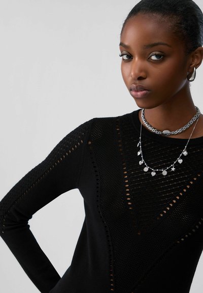 Jeune femme portant un haut noir texturé à manches longues, un collier chaîne argenté avec des breloques, et des boucles d'oreilles argentées assorties, sur fond uni.