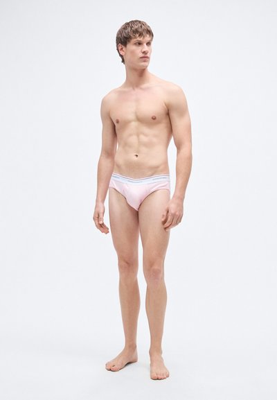 Ροζ βαμβακερά σλιπ με ελαστική μέση και αντίθετο κείμενο. Σώμα που εφαρμόζει τέλεια και λεία υφή. Το μοντέλο στέκεται ξυπόλυτο.