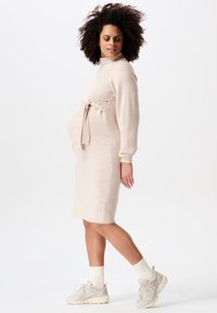 Gebreide, lichtbeige jurk met een hoge hals, een ceintuur in de taille en lange mouwen, gecombineerd met lichte sneakers en witte sokken. Gladde textuur.