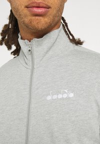 Homme à la peau brun clair et aux dreadlocks, portant une veste zippée gris clair avec un logo Diadora blanc sur la poitrine.