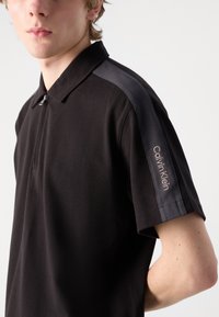 Čierna polo košeľa so zipsom v krku, s šedými akcentmi na pleciach a s vyšitým nápisom "Calvin Klein" na ľavom rukáve. Textúrovaná tkanina.