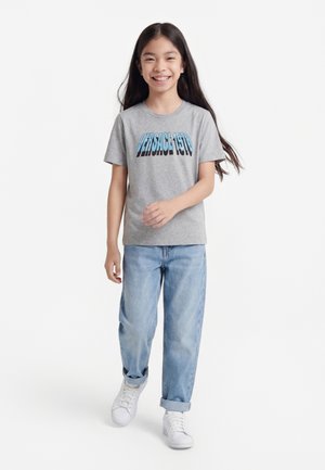 Smilende pige går fremad iført en grå "Versace 1978" t-shirt, lyseblå jeans med opsmøg og hvide sneakers på hvid baggrund.