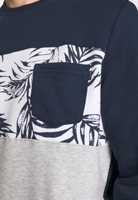 Sudadera en navy y gris con un panel blanco con un patrón de hojas y un pequeño bolsillo delantero. Enfocada en un estilo casual y un diseño moderno.