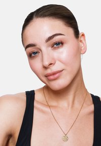 Collier en pendentif en or avec un charme circulaire texturé, suspendu à une chaîne délicate, porté sur un haut noir. La peau a une apparence lisse.