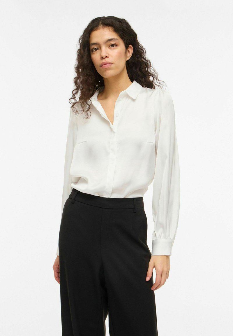 Žena s kudrnatými tmavými vlasy, která má na sobě bílou blůzu s dlouhým rukávem zastrčenou do černých kalhot s vysokým pasem, stojící před bílým pozadím.