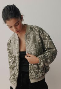Short-sequence bomber jacket featuring silver sequins with floral patterns, a light gray ribbed collar, and cuffs, and a fitted silhouette.