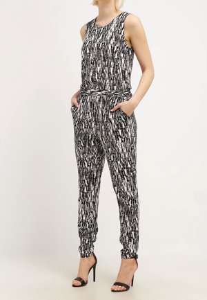 Kobieta w czarno-białym, wzorzystym kombinezonie bez rękawów o zwężanych nogawkach i czarnych sandałach na wysokim obcasie, ręce w kieszeniach.