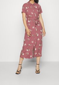 Różowa sukienka z krótkim rękawem, w kwiatowy wzór, z zapięciem na guziki z przodu i wiązanym w pasie. Średniej długości, o prostym kroju. Czarne sandały na obcasie.