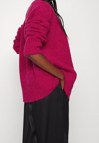 Fuchsiový oversize svetr s texturovaným pletením, ohrnutými manžetami a bočními rozparky, v kombinaci s černou skladanou sukní pro kontrast.