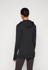 Melna garām piedurknēm kapuce ar pieguļošu dizainu un mīkstu tekstūru. Iespējo saules kabatu un regulējamu kapuci. Nēsāta ar melnajām legingām.