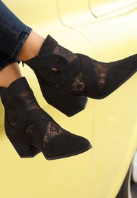 Melnas potītes zābakus ar smailu purngalu, ar mežģīņu paneļiem, mīkstu zamšādu un zemu bloku papēdi. Tekstūras akcenti uzlabo dizainu.