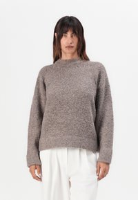 Pull en maille marron avec une finition texturée, à manches longues et col haut. Associé à un pantalon blanc, créant une silhouette décontractée.