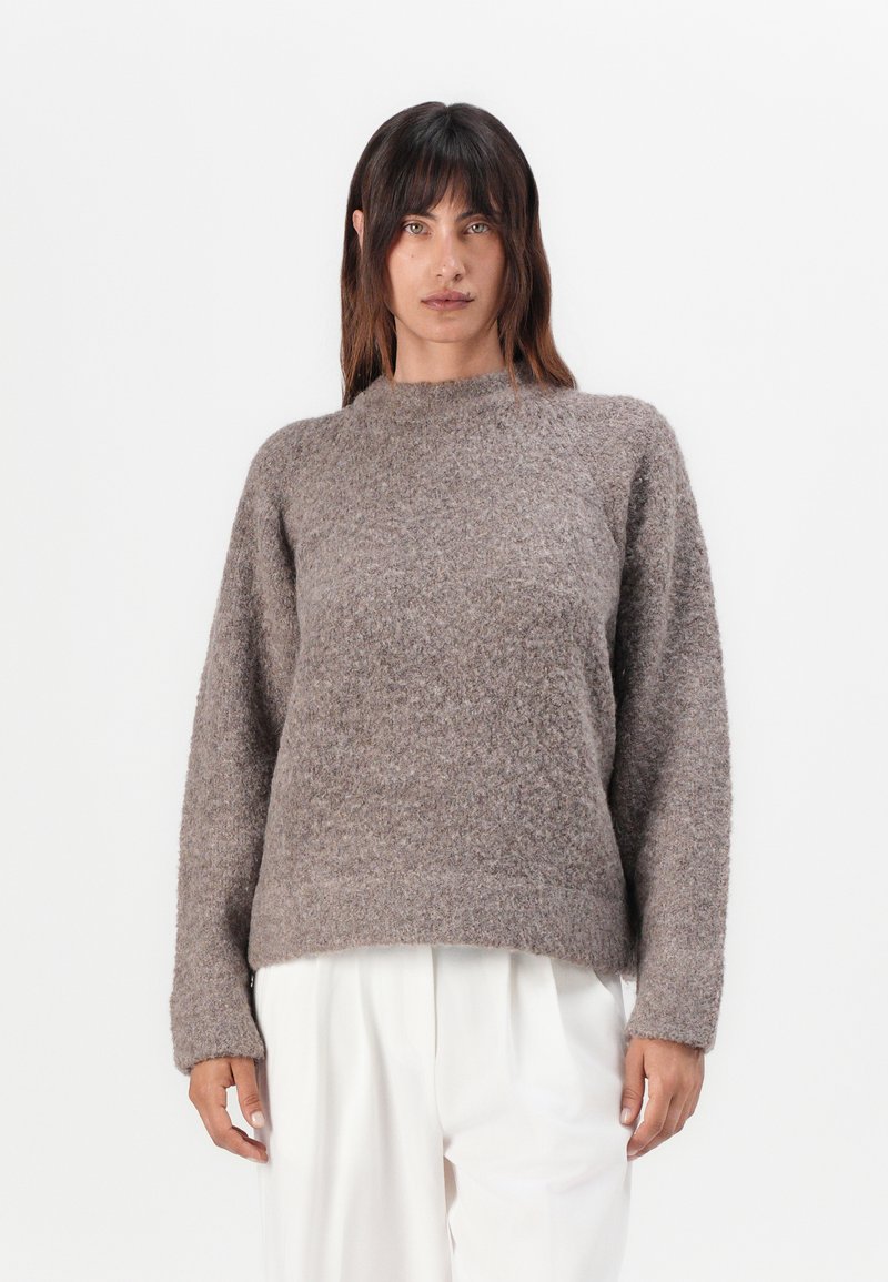 Pull en maille marron avec une finition texturée, à manches longues et col haut. Associé à un pantalon blanc, créant une silhouette décontractée.