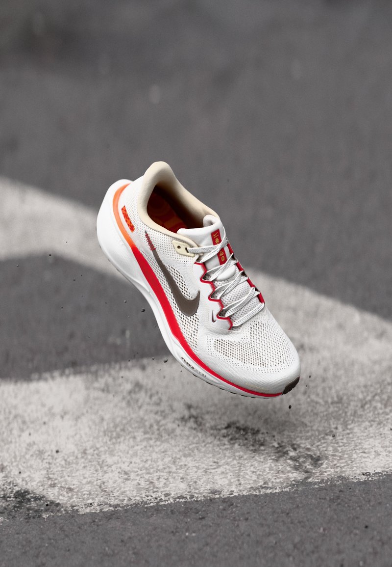 Valge spordijalgpall, millel on punased ja oranžid aktsendid, võrgust pealmine osa, pehmendatud krae ja peegeldav Nike swoosh, näib hõljuvat halli pinna kohal.