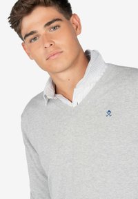 Suéter gris de cuello en V hecho de una tela suave, con un pequeño logo azul en el pecho, superpuesto a una camisa de cuello blanca y azul a rayas.