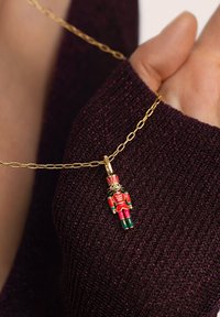 Naszyjnik z łańcuszkiem ze złota z zawieszką w kształcie dziadka do orzechów w kolorach czerwonym, zielonym i złotym. W tle widoczny jest teksturowany, ciemnofioletowy sweter.