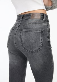 Jeans de mezclilla gris de cintura alta con un diseño ajustado. Incluye dos bolsillos traseros y un parche de logo marrón en la cintura. Textura suave.