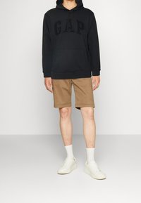 Person som bär en svart GAP-hoodie, beige knälånga shorts, vita strumpor och vita sneakers som står mot en enkel bakgrund.